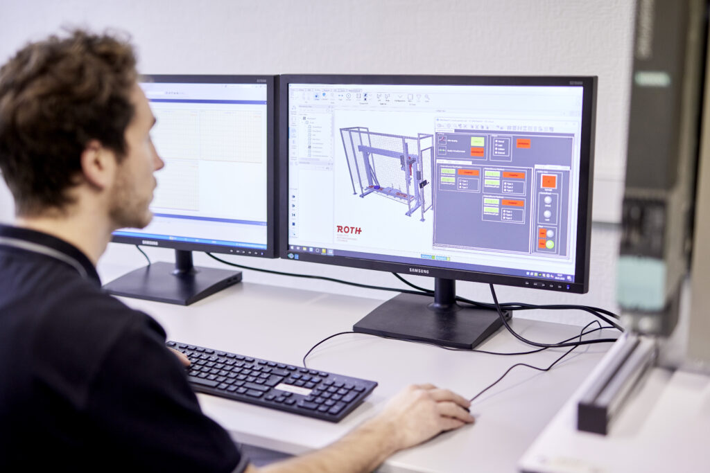 Ein junger Mann sitzt vor zwei Monitoren und führt die virtuelle Inbetriebnahme einer technischen Anlage durch. Auf den Bildschirmen sind schematische Darstellungen und Steuerungsoberflächen zu sehen.