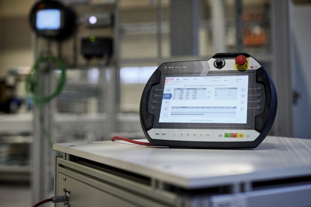 Ein modernes Handbediengerät im Vordergrund, im unscharfen Hintergrund ist eine industrielle Anlage mit Schutzzaun zu sehen.