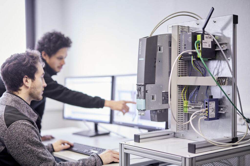 Im Vordergrund zu sehen ein Steuerungsaufbau und dahinter 2 Männer die am Bildschirm deine simulierte Anlage begutachten.