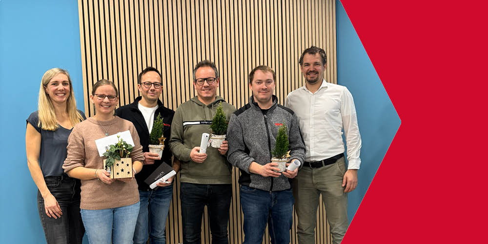 Zwei Frauen und vier Männer stehen nebeneinander vor einer Wand und halten Präsente anlässlich ihres Firmenjubiläums in den Händen.