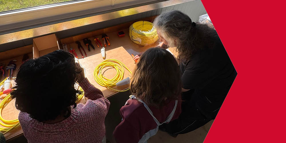 Eine Frau und 2 Mädchen arbeiten an einer Werkbank vor ihnen liegen Verlängerungskabel und verschiedenes Werkzeug.