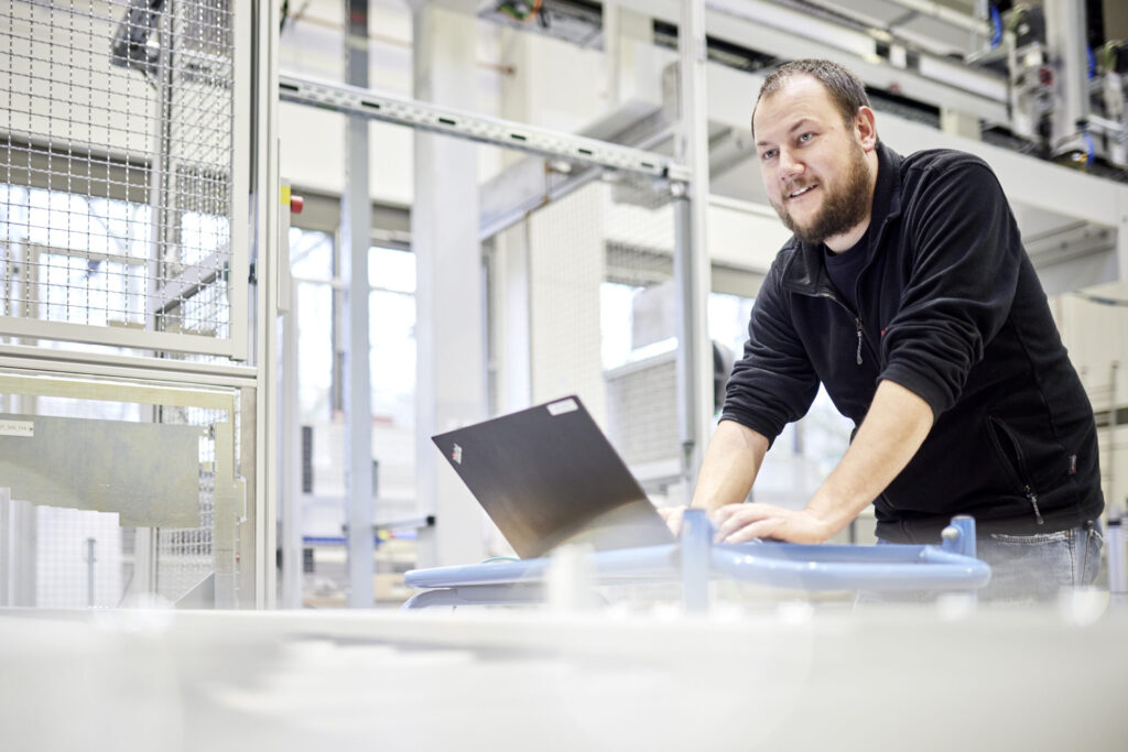 Ein Mann arbeitet an einem Laptop. Sein Blick ist auf einen nicht sichtbaren Punkt vor ihm gerichtet, während im Hintergrund eine moderne Industrieanlage mit Schutzzaun und Ladeportal zu sehen ist.