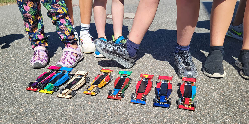 Viele Kinderfüße stehen hinter einer Reihe bunter, solarbetriebener Rennautos.